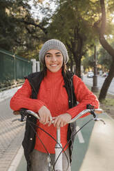 Junge Frau in warmer Kleidung steht mit Fahrrad auf der Straße - JRVF02514