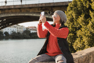 Lächelnde junge Frau beim Fotografieren mit dem Smartphone an einem sonnigen Tag - JRVF02517