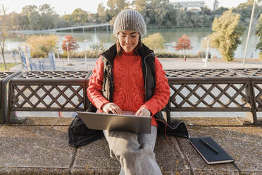 Young working woman using laptop sitting with diary on bench - JRVF02524