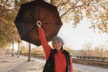 Lächelnde junge Frau mit Regenschirm auf dem Fußweg stehend - JRVF02526