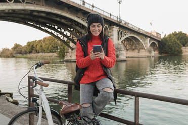 Smiling young woman with bicycle using smart phone sitting on railing - JRVF02531