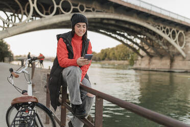 Young woman using mobile phone sitting on railing by bicycle at riverbank - JRVF02532