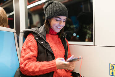 Lächelnde junge Frau, die ein Smartphone benutzt und Musik über In-Ear-Kopfhörer in der Straßenbahn hört - JRVF02538