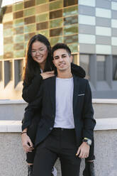 Businessman leaning on businesswoman sitting on wall in front of office building - JRVF02568