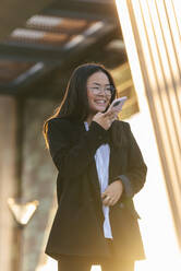 Lächelnde Geschäftsfrau im Gespräch über Lautsprecher vor einem Gebäude - JRVF02573