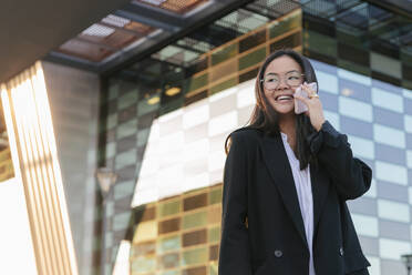 Junge Geschäftsfrau, die vor einem Bürogebäude mit einem Mobiltelefon spricht - JRVF02575