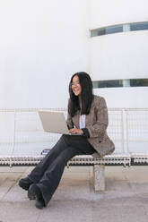 Lächelnde Geschäftsfrau mit Laptop auf einer Bank sitzend - JRVF02595