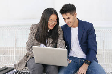 Smiling businesswoman showing laptop to colleague sitting on bench - JRVF02598