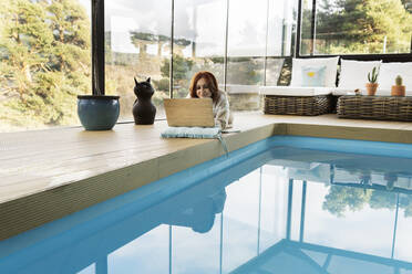 Frau benutzt Laptop am Swimmingpool in einem modernen Haus - JCCMF05201