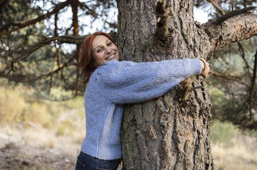 Smiling woman embracing tree trunk in park - JCCMF05222