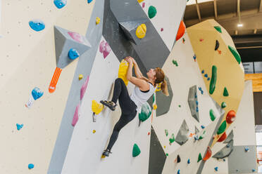Ganzkörper-Seitenansicht einer aktiven Bergsteigerin in Sportkleidung an einer künstlichen Kletterwand mit bunten Griffen beim Training in einer modernen Sporthalle - ADSF33185