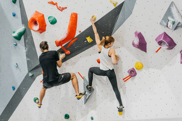 Rückenansicht eines sportlichen Paares in Sportkleidung, das beim gemeinsamen Training in einem hellen, modernen Fitnessstudio eine Kunstwand mit bunten Griffen erklimmt - ADSF33189