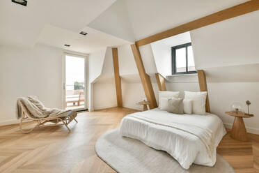 Interior of spacious light contemporary bedroom furnished with comfortable bed placed on parquet floor near white sloped uneven wall with decorative beams and window - ADSF33222