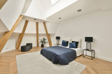 Interior of contemporary spacious light bedroom furnished with comfortable bed placed on carpet on parquet floor near white sloped walls with decorative wooden beams - ADSF33223