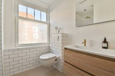 Weiße Keramiktoilette an weißer Backsteinwand neben Holzschrank mit Waschbecken in hellem, modernem Badezimmer mit Fenster und Spiegel - ADSF33235
