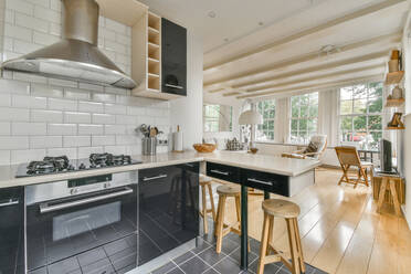 Counter with gas stove with oven placed at tiled wall in stylish kitchen with modern appliances and cabinets next to a worktop with wooden chairs and at the back a spacious living room with large windows - ADSF33240