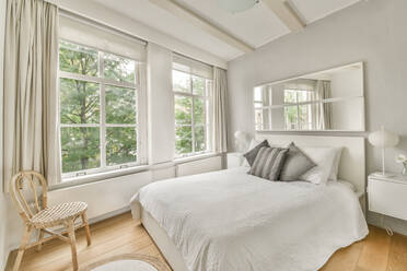 Bett mit weißer Decke und Kissen an der Wand mit Spiegel neben Korbstuhl in stilvollem Schlafzimmer mit Fenstern mit Blick auf grüne Bäume platziert - ADSF33241