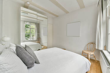 Bett mit weißer Decke und Kissen bedeckt neben dem Spiegelschrank in der Nähe von Korbstuhl in stilvollem Schlafzimmer mit Fenstern mit Blick auf grüne Bäume platziert - ADSF33242