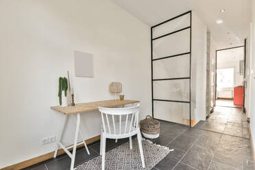 Weißer Stuhl am Tisch mit Dekorationen in der Nähe von Weidenkorb und Fenster in hellem Raum mit Glastür in stilvoller Wohnung - ADSF33275