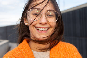 Junge positive Frau mit Brille und orangefarbener Strickjacke, die entspannt in die Kamera schaut und mit den Haaren wedelt - ADSF33359