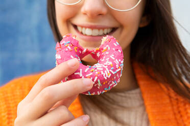 Positive cropped unrecognizable woman in knitted warm cardigan eating tasty pink sprinkled donut - ADSF33360