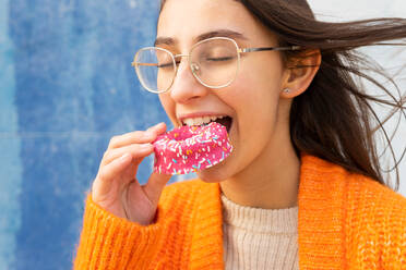 Positive young woman in eyeglasses and knitted warm cardigan eating tasty pink sprinkled donut with closed eyes - ADSF33361