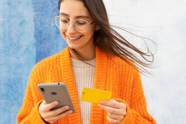 Von unten: fröhliche Frau mit Brille und orangefarbener Strickjacke, die mit einer Plastikkarte auf einem Smartphone für einen Online-Einkauf bezahlt - ADSF33362
