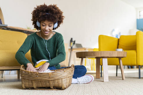 Junge Afro-Frau, die im Schneidersitz neben einem Korb im Wohnzimmer sitzt und Musik hört - GIOF14666