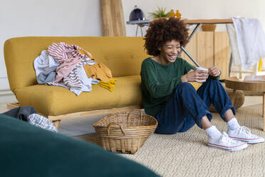 Afro woman holding coffee mug laughing by basket in living room - GIOF14670