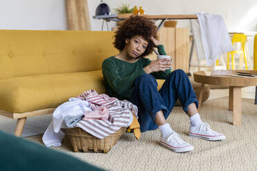 Tired Afro woman with hand in hair holding coffee mug sitting by basket at home - GIOF14671