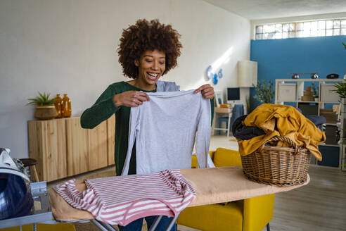 Glückliche Frau hält T-Shirt am Bügelbrett im Wohnzimmer - GIOF14679