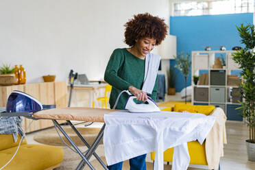 Smiling young Afro woman ironing shirt on board in living room - GIOF14688