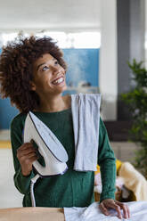 Verspielte junge Afro-Frau, die ein Bügeleisen mit einem Hemd auf einem Brett im Wohnzimmer hält - GIOF14691
