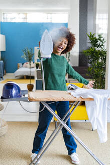 Fröhliche junge Afro-Frau, die Dampf aus einem Bügeleisen bläst, das an einem Brett mit einem Hemd im Wohnzimmer steht - GIOF14695