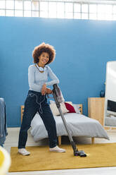 Lächelnde junge Afro-Frau, die mit einem Staubsauger auf einem Teppich im Schlafzimmer steht - GIOF14699