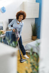 Lächelnde junge Afro-Frau reinigt Teppich mit Staubsauger im Schlafzimmer zu Hause - GIOF14701