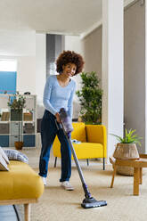 Lächelnde junge Afro-Frau beim Reinigen des Teppichs im Wohnzimmer zu Hause - GIOF14706