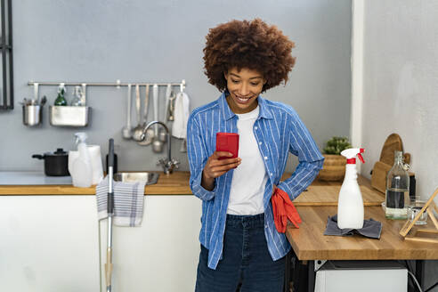 Lächelnde junge Afro-Frau, die mit einer Sprühflasche an der Küchentheke eine Textnachricht auf einem Smartphone schreibt - GIOF14718