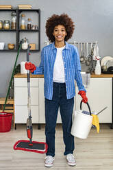 Smiling young Afro woman holding cleaning bucket and mop in kitchen - GIOF14721