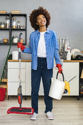 Thoughtful young Afro woman holding cleaning bucket and mop in kitchen - GIOF14722