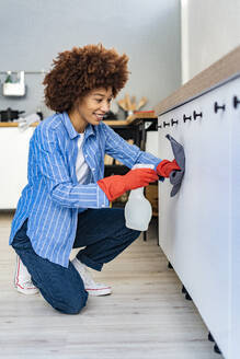 Lächelnde Afro-Frau kniet und sprüht Desinfektion auf Schrank in der Küche - GIOF14723