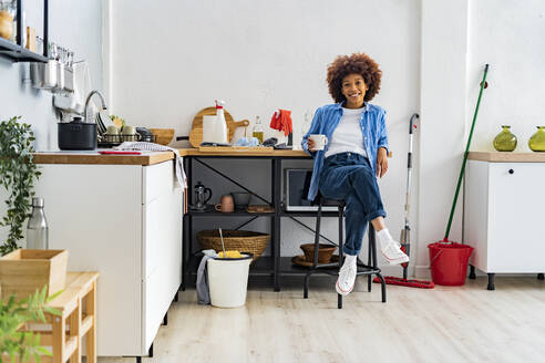 Lächelnde junge Afro-Frau, die auf einem Hocker sitzt und sich auf die Küchentheke stützt, um sich zu Hause zu entspannen - GIOF14731