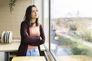 Young woman with arms crossed looking out of window at home - XLGF02520