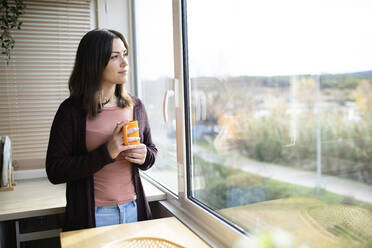Woman with mug looking out of window standing at home - XLGF02522