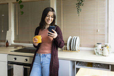 Smiling woman using smart phone holding mug leaning on kitchen counter at home - XLGF02525