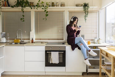 Frau mit Kopfhörern und Smartphone auf dem Küchentisch zu Hause - XLGF02533