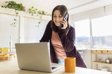 Young businesswoman talking on smart phone by laptop in kitchen at home - XLGF02553