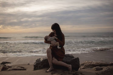 Caring mother holding newborn son sitting on rock at beach - GMLF01215