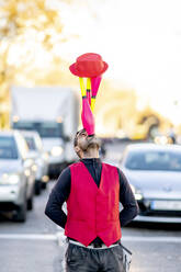 Performer balancing juggling pins and hat on nose in front of cars - OCMF02316