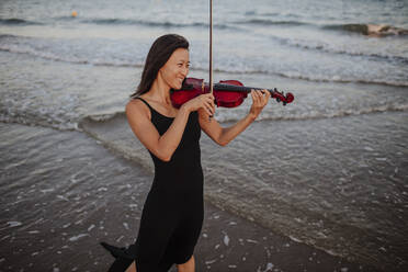 Happy woman playing violin in sea at beach - GMLF01228